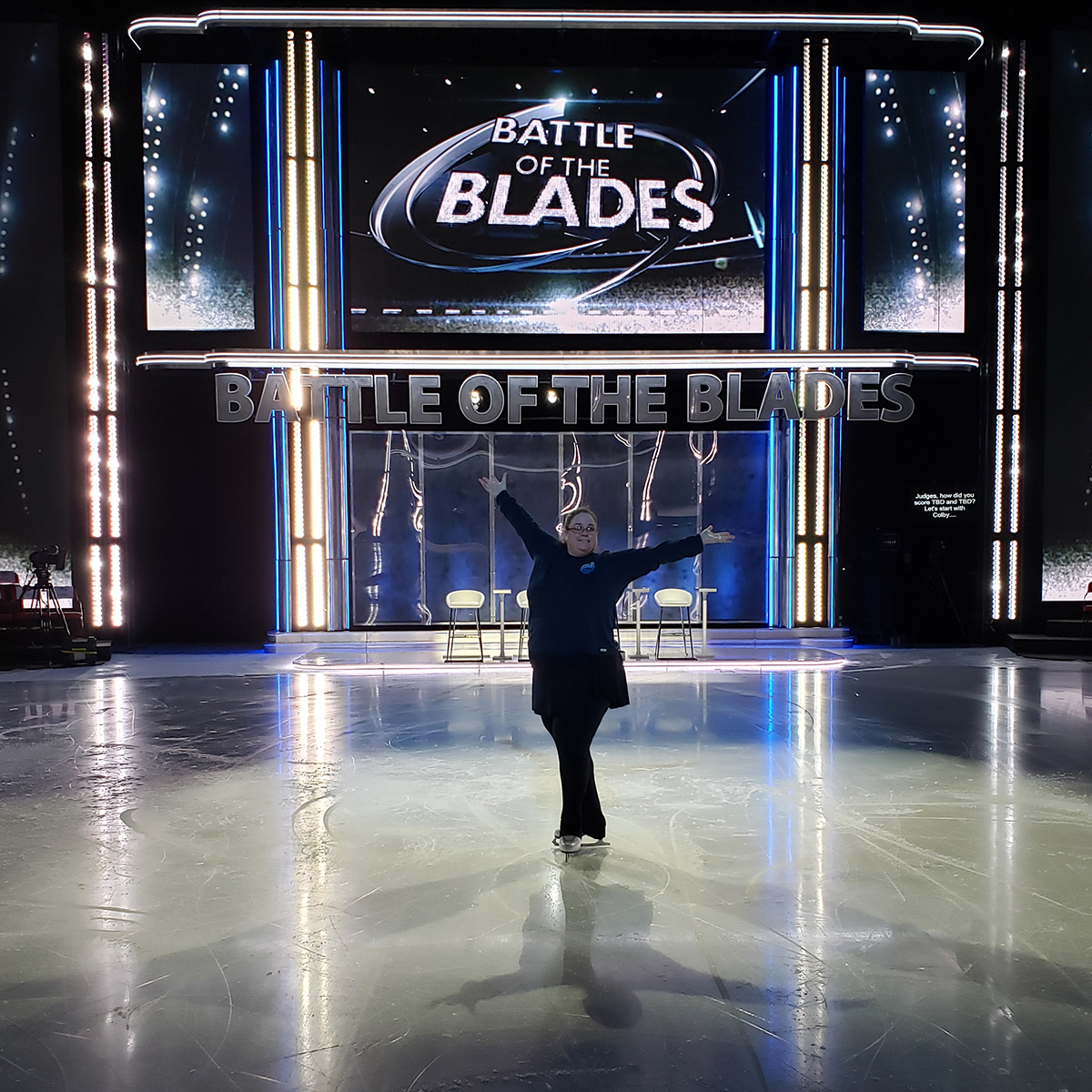A large middle aged figure skater in front of the Battle of the Blades backdrop.