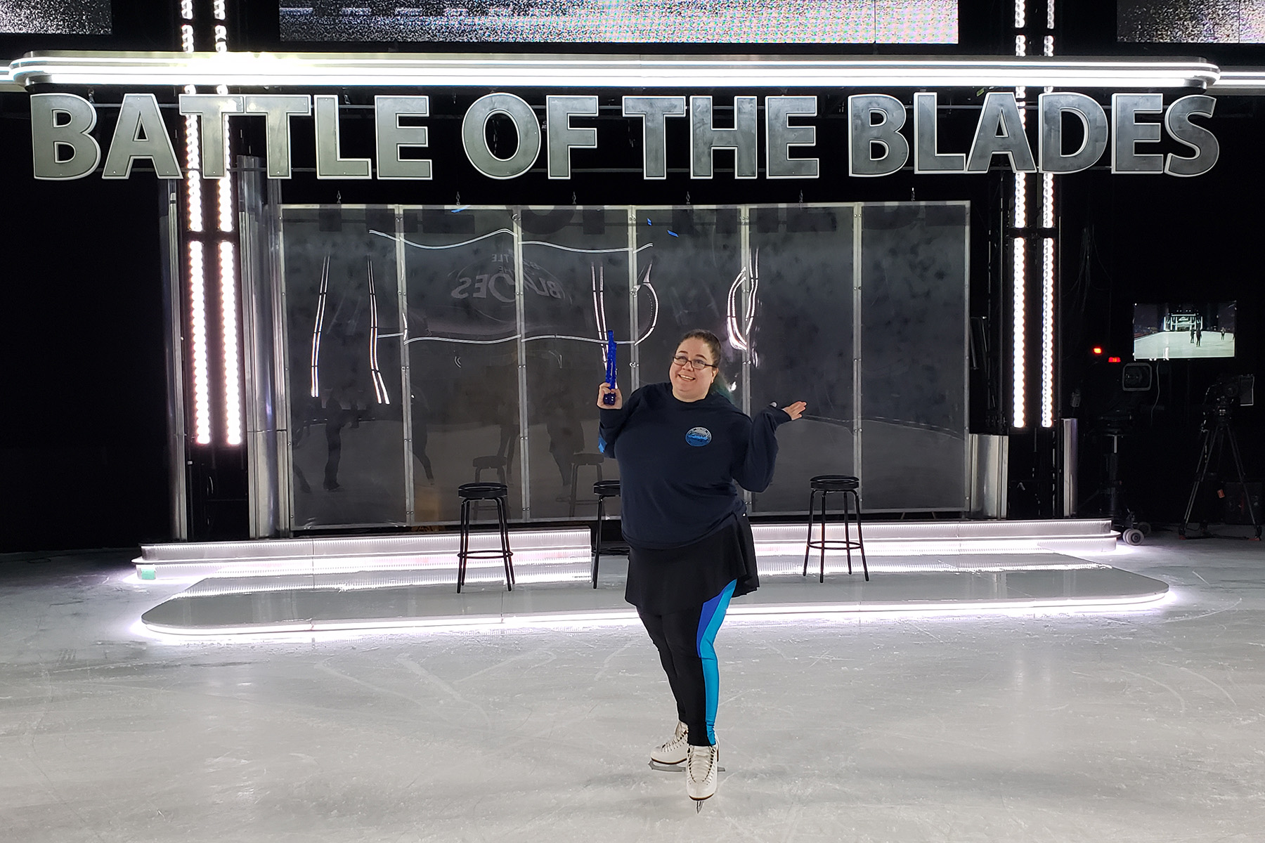 A large middle aged figure skater in front of the Battle of the Blades backdrop.