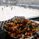 A plate of buffalo chicken poutine held up with an ice rink and hockey game in the background.