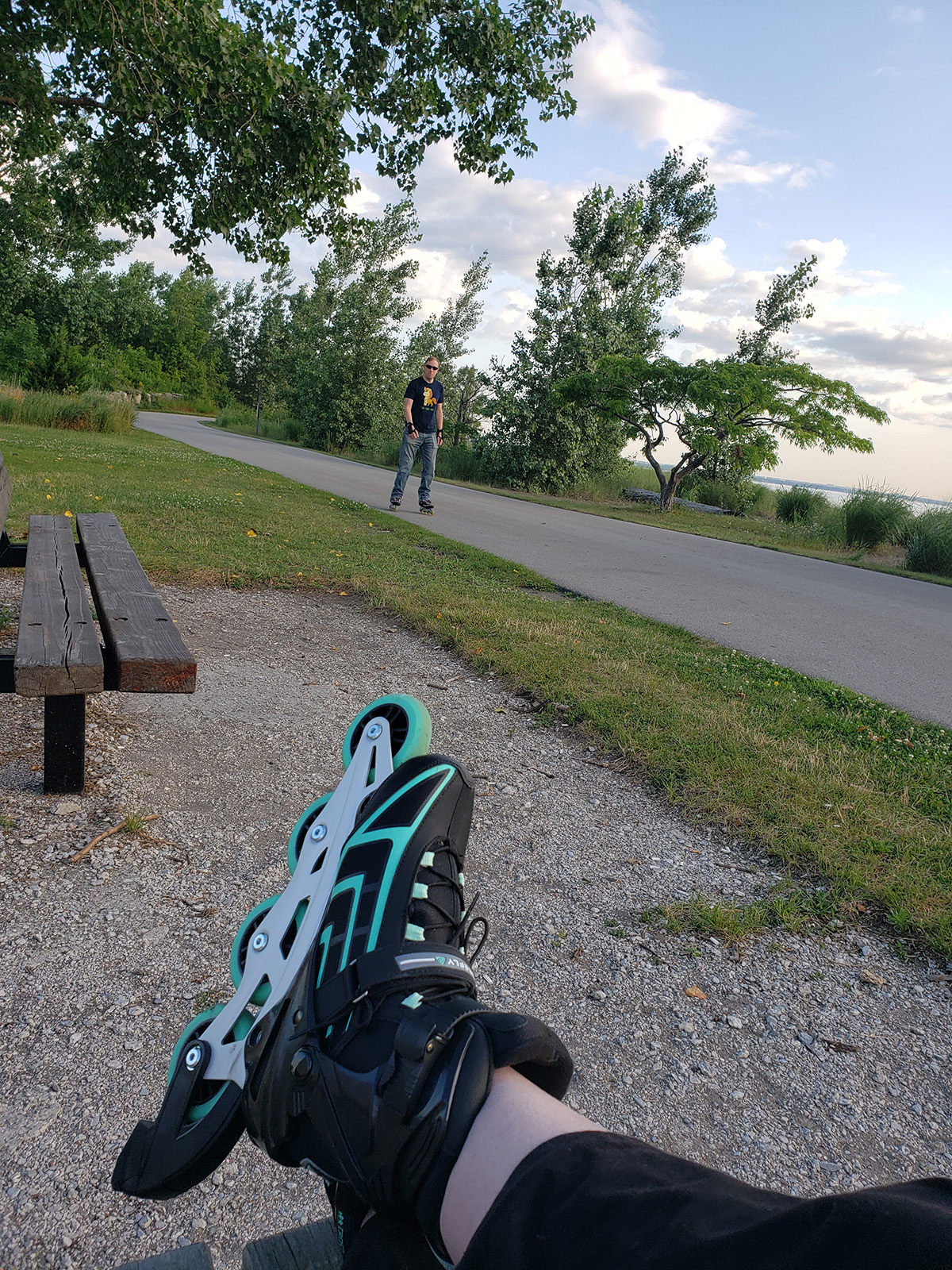 Legs wearing inline skates are crossed, with a man skating ona  trail in the background.