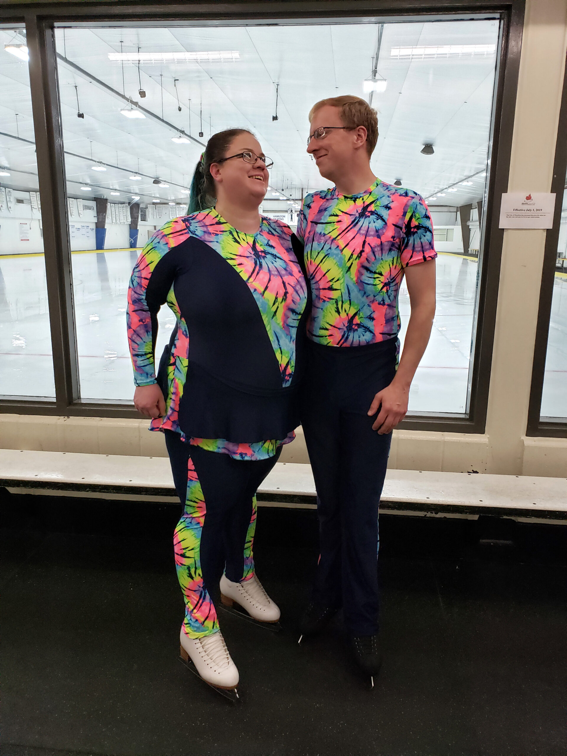 A middle aged couple in bright neon tie-dye skating costumes smiling at each other with a rink in the background.