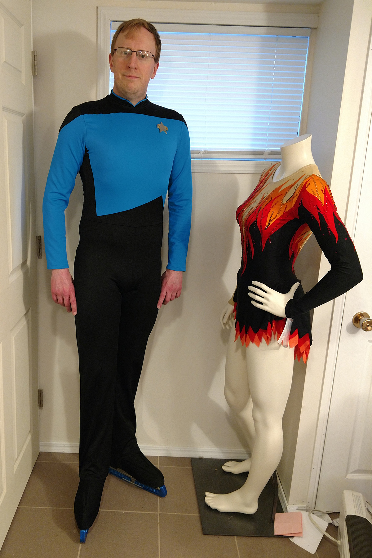 A blond man wearing a 1 piece figure skating costume that looks like a Star Trek The Next Generation science officer uniform.