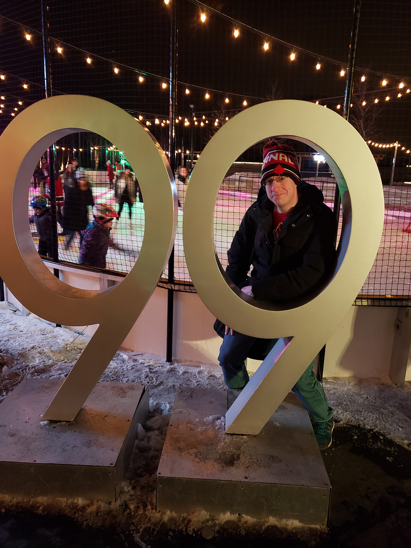 A blond man standing behind a large 99 in front of a skating rink.